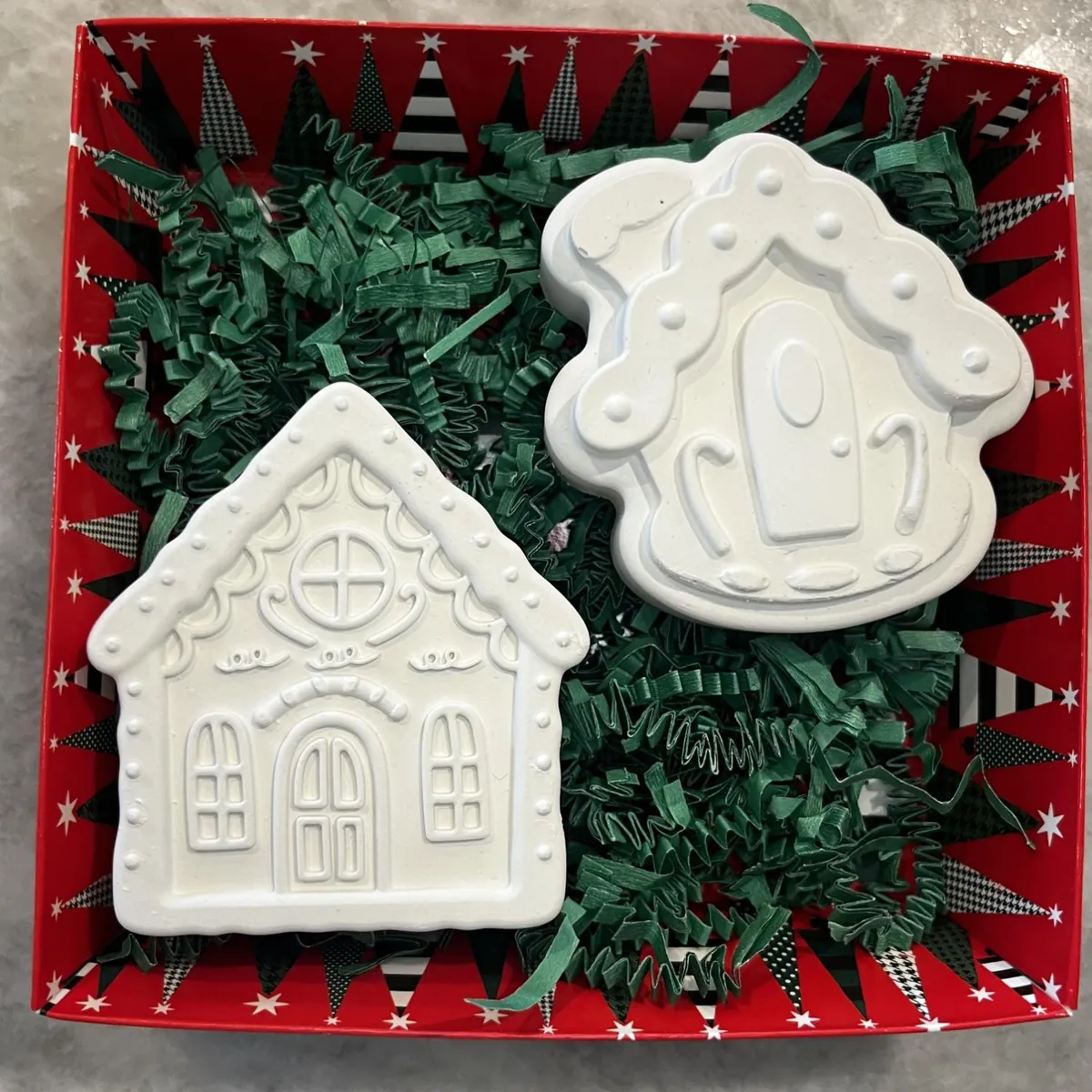 Two white plaster gingerbread houses in a red holiday box with green shred